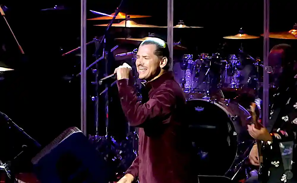 A man in a maroon shirt sings into a microphone on a stage with drums behind him.