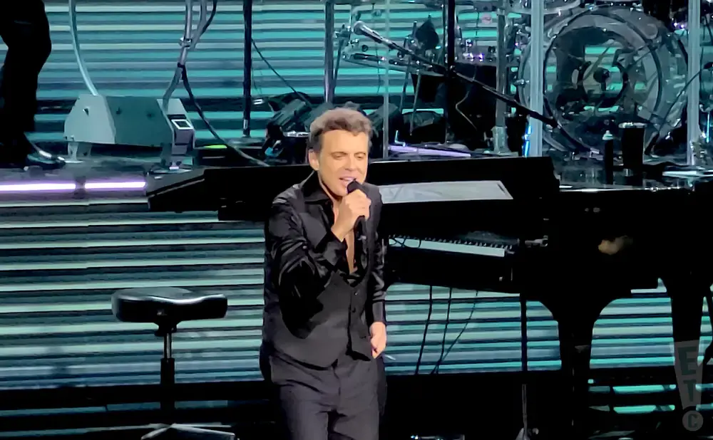 A singer in a black shirt and pants performs on stage, holding a microphone.
