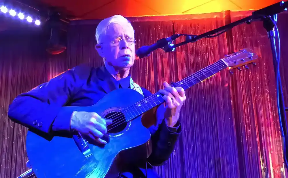 Man with white hair and glasses plays an acoustic guitar on stage under blue and pink lights.