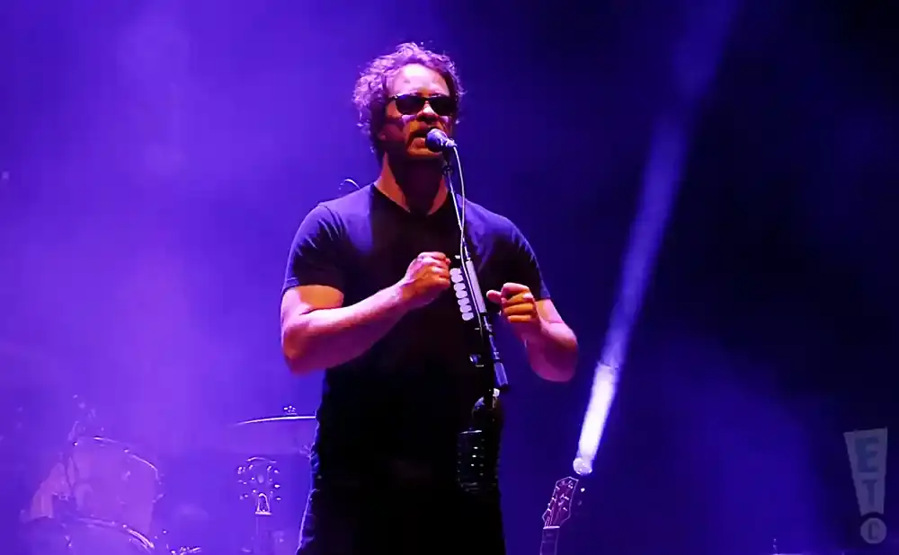 A musician with sunglasses sings into a microphone while holding a pipe-like instrument.
