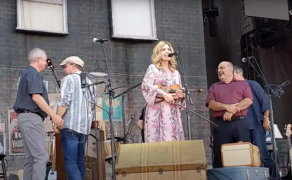 A woman in a floral dress plays a violin while a man in a maroon shirt smiles on stage.