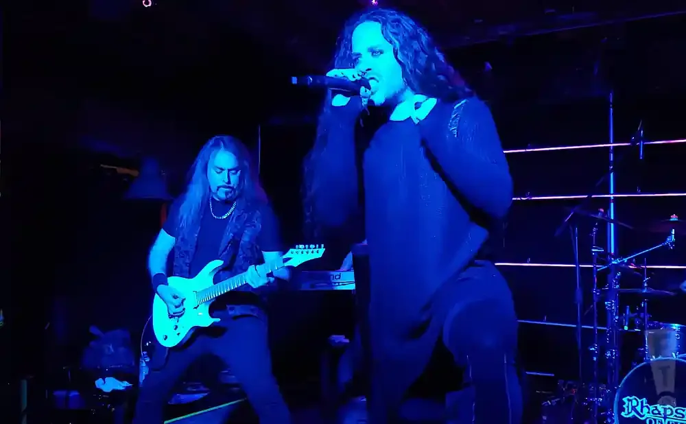 A rock singer holds a microphone on stage with a guitarist and drums in the background.