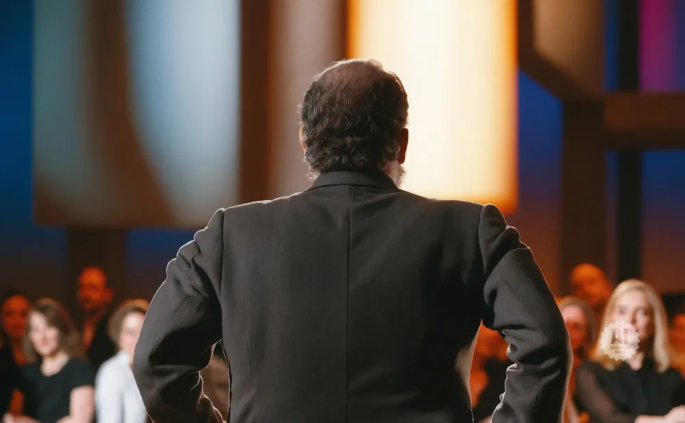 Man in a suit facing away from the camera, speaking to an audience.