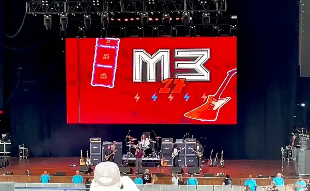 A rock band performs on stage with a large M3 logo and guitar graphics on a red screen backdrop.