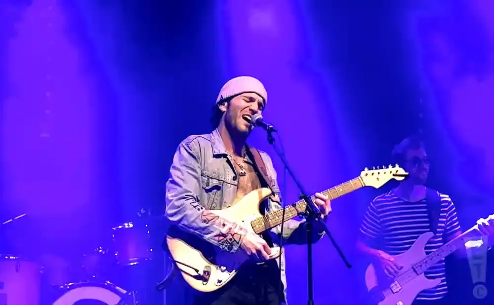 A male singer in a denim jacket and beanie plays a guitar and sings into a microphone on a stage with blue lighting.