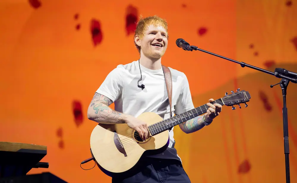 Ed Sheeran plays an acoustic guitar on stage with an orange backdrop.