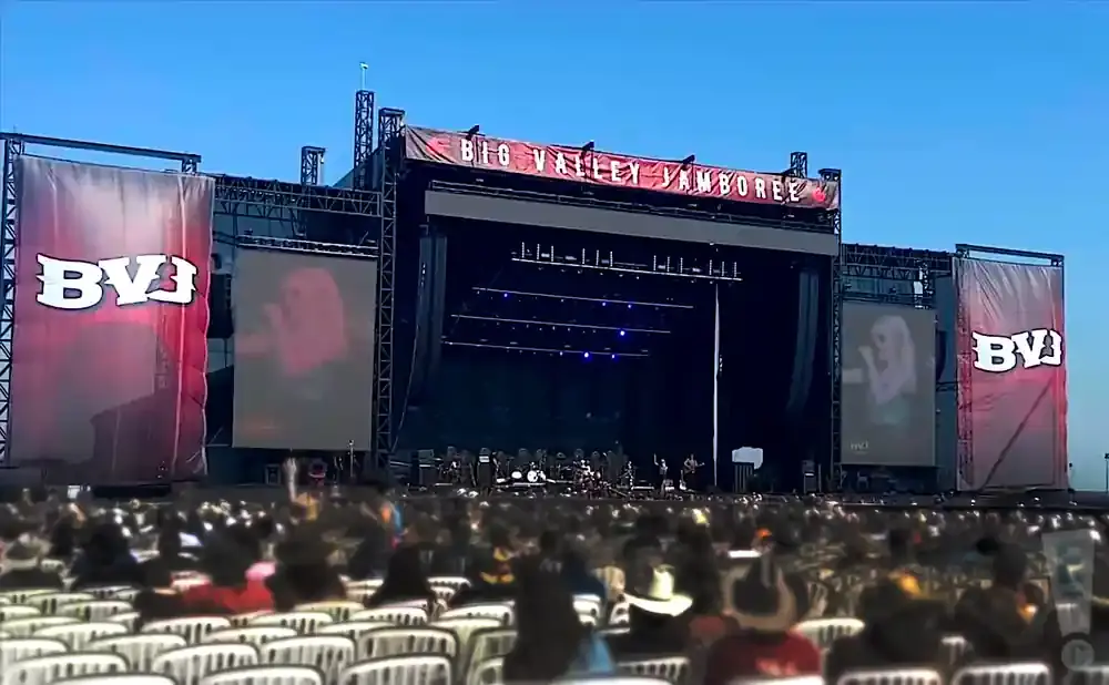 A large outdoor concert stage with screens showing performers, banners, and a crowd in the foreground.