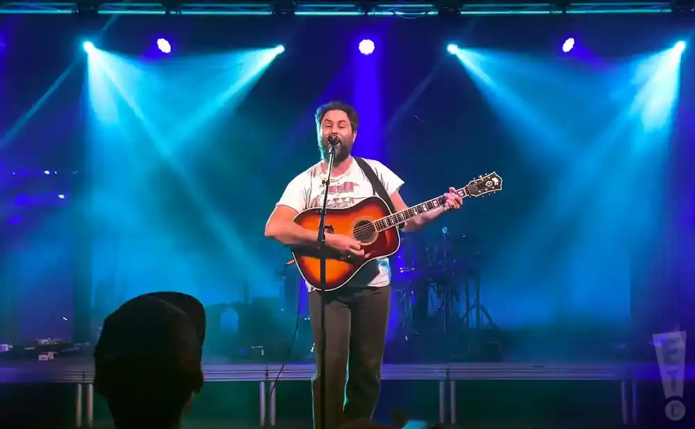 Musician with beard plays acoustic guitar and sings on stage with blue spotlights.