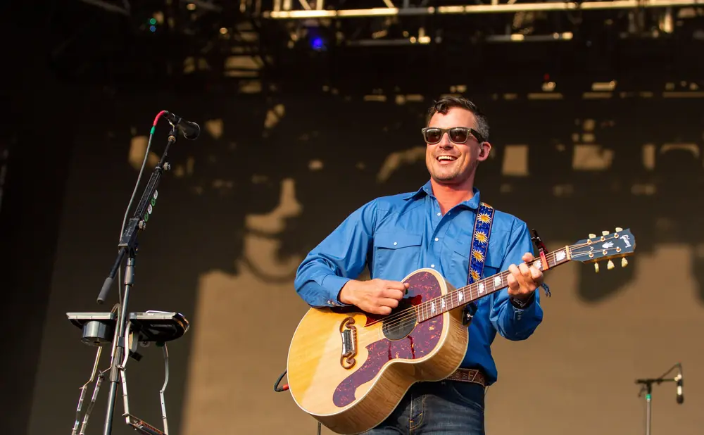 A man in sunglasses and a blue shirt plays an acoustic guitar on stage.