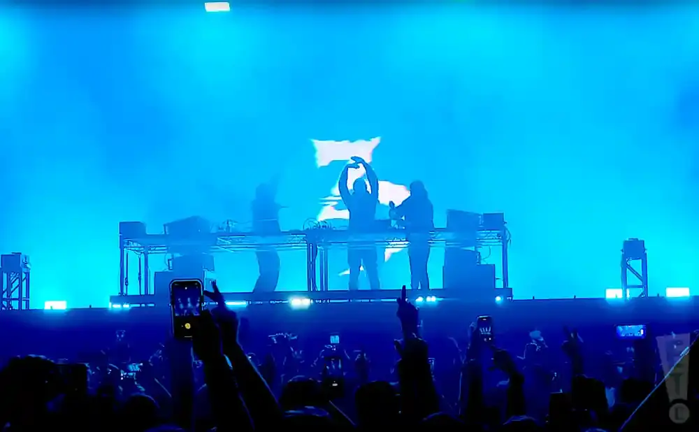 A DJ trio performs on a stage bathed in blue light, with a crowd in the foreground holding up phones.