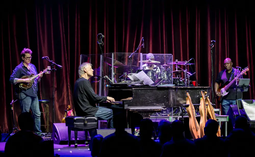 Musicians perform on a stage with a piano, drums, bass, and mandolin.