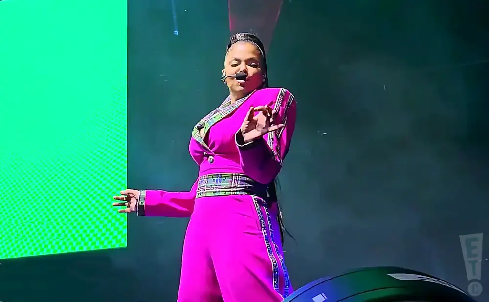 A woman in a bright pink jumpsuit and jacket with plaid trim sings on stage, gesturing with her hand.