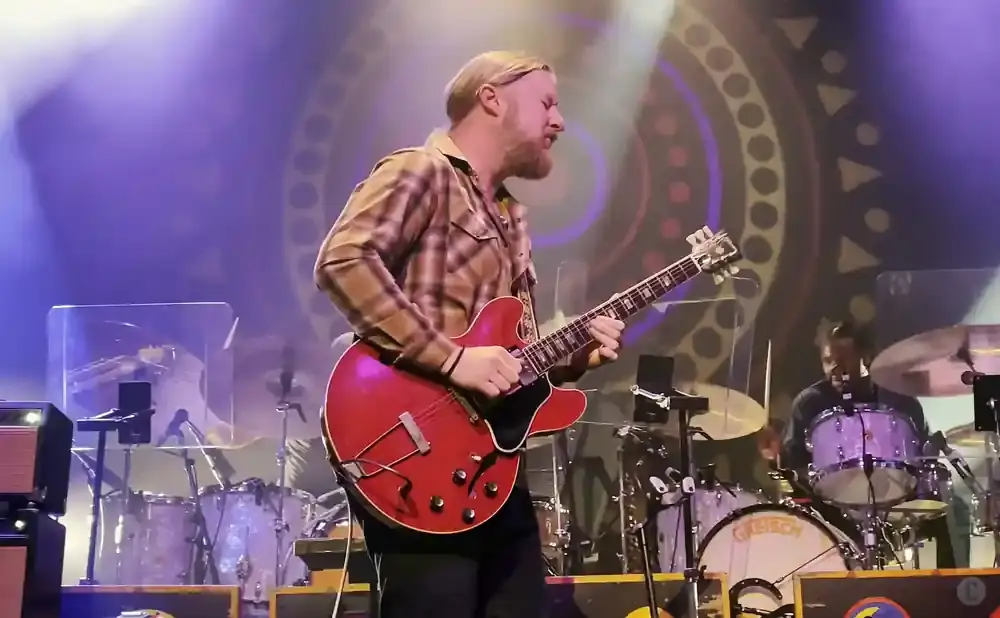 A musician plays a red electric guitar on stage, with a drummer in the background.