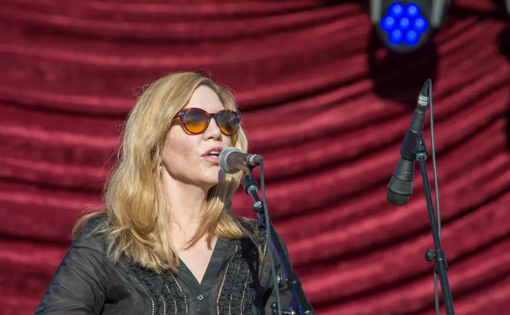 A blonde woman in sunglasses sings into a microphone on a stage with a red velvet backdrop.