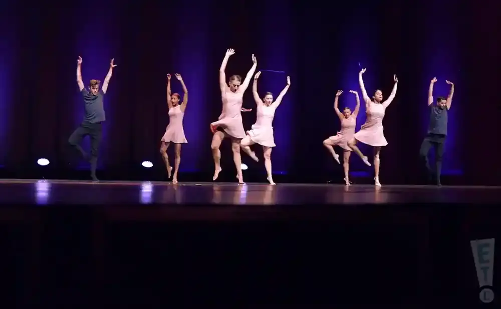 Dancers in pink dresses and one man in a grey shirt and black pants perform on a stage with a purple backdrop.
