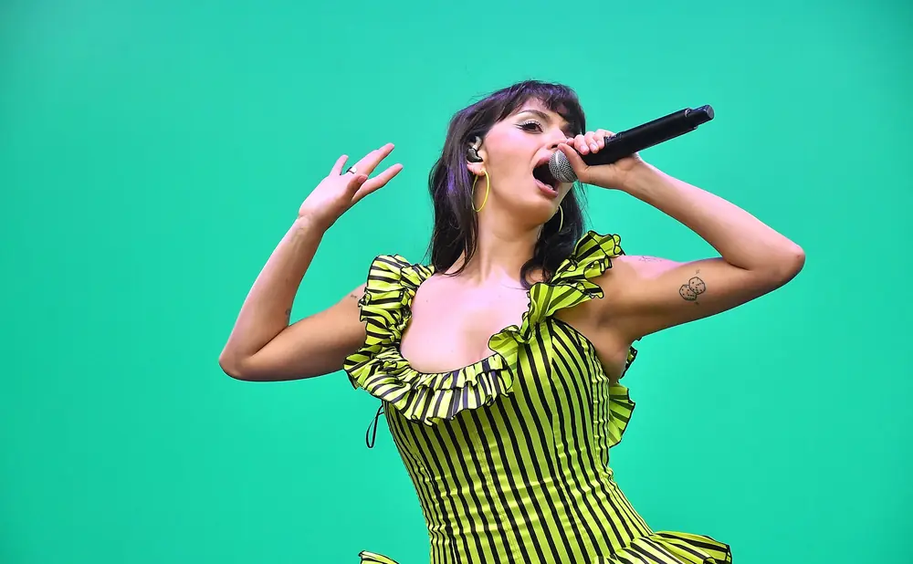 Singer with purple-streaked hair in a neon green and black striped dress sings into a microphone.