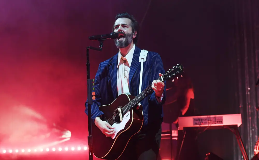 A man with a beard and a blue pinstripe suit sings into a microphone while playing an acoustic guitar on stage.