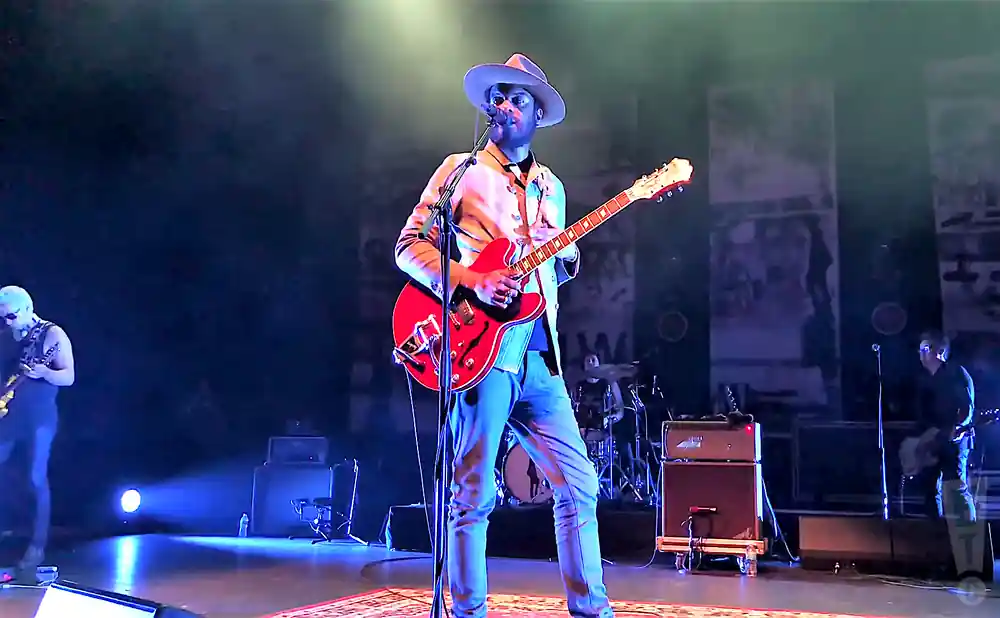 A man in a hat plays a red electric guitar on a dimly lit stage.