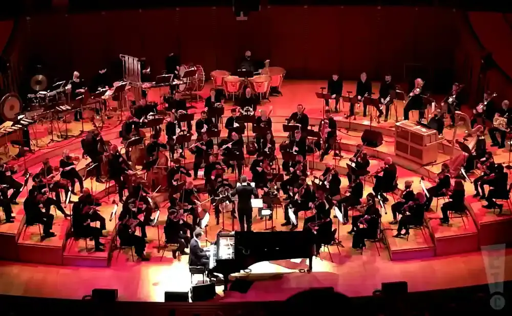 An orchestra plays on a tiered stage under red lighting, with a conductor leading from the front.