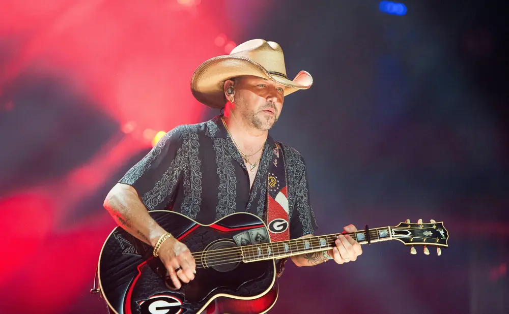 A man in a cowboy hat plays an acoustic guitar on stage with red and blue lights.
