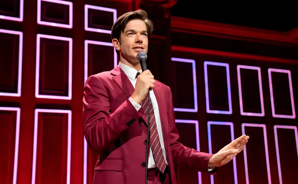 Comedian John Mulaney in a red suit on stage with a microphone, speaking to an audience.