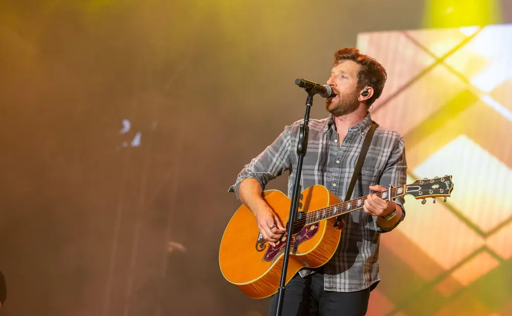 A male singer with reddish-brown hair, wearing a plaid shirt, sings into a microphone while playing an acoustic guitar on stage.