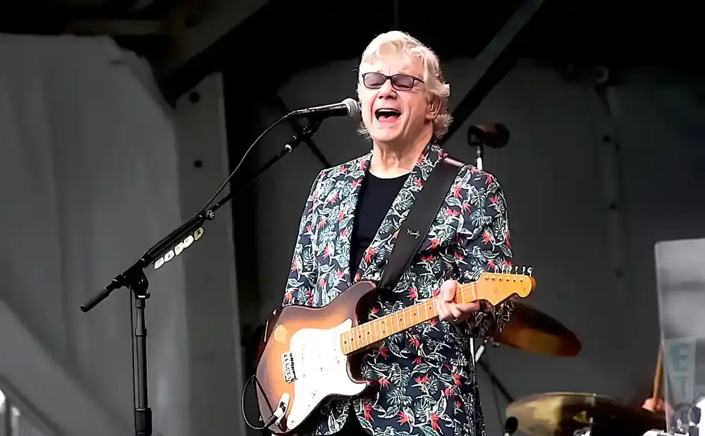 A man in a floral jacket sings into a microphone while playing a guitar.