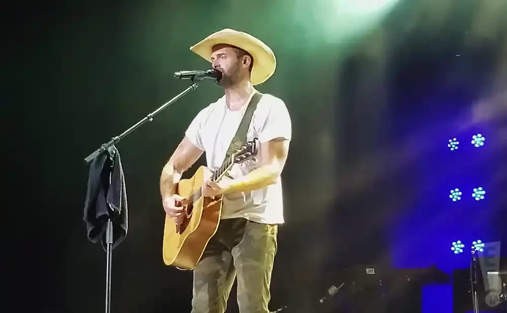 A male singer in a cowboy hat plays an acoustic guitar and sings into a microphone on a dark stage with blue lights.