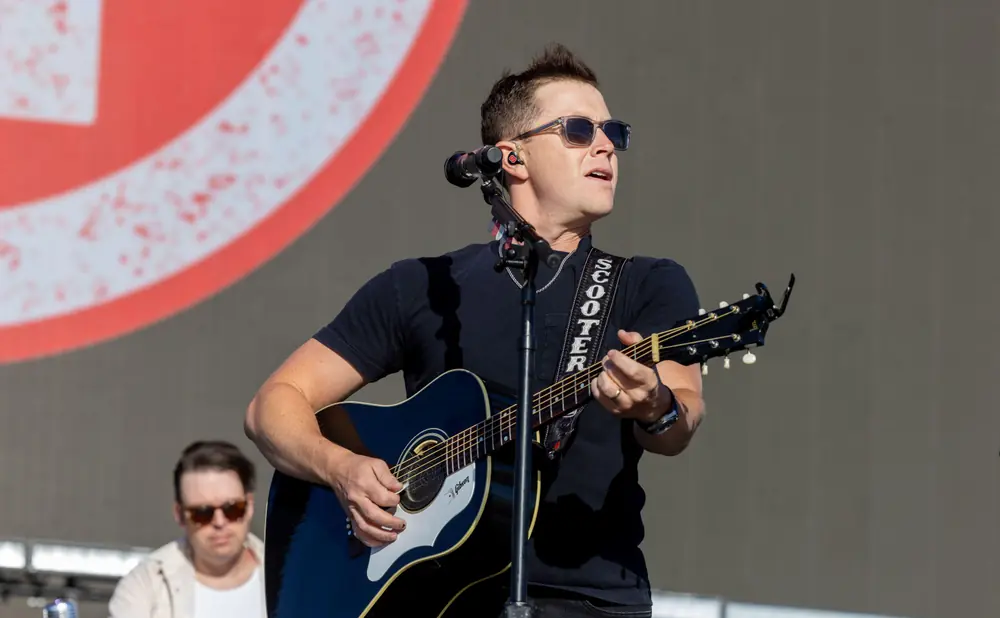 A man with sunglasses plays an acoustic guitar on stage, with another man blurred in the background.