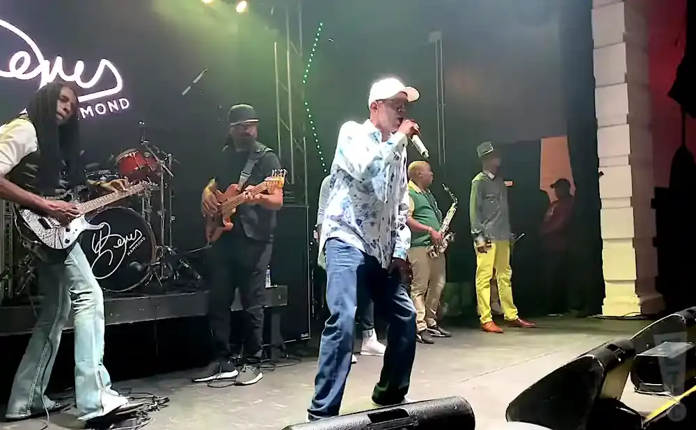A reggae band performs on stage with a lead singer, guitarist, bassist, drummer, and saxophonist.