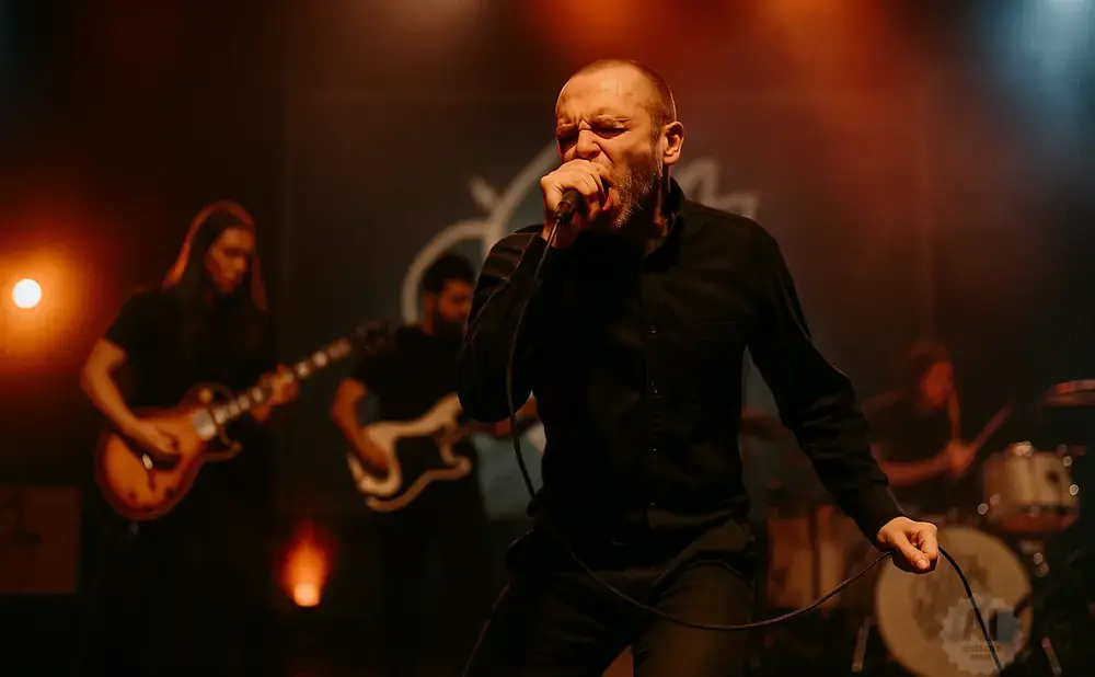 A male singer in a black shirt sings into a microphone on stage, with bandmates playing guitar and drums behind him.