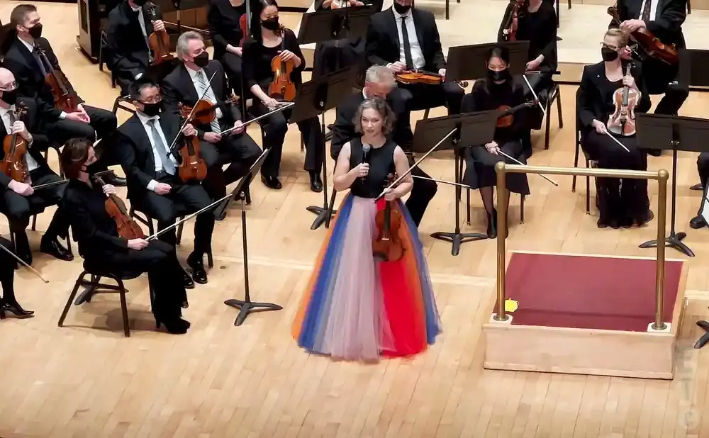 A conductor in a colorful skirt stands with a violin in front of an orchestra.