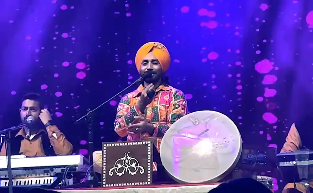 Musicians performing on stage with keyboard, drum, and traditional Indian instrument, under purple stage lighting.