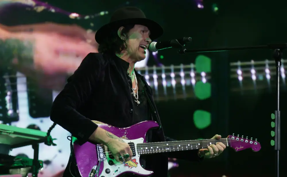 A man in a hat plays a purple electric guitar on stage, singing into a microphone.