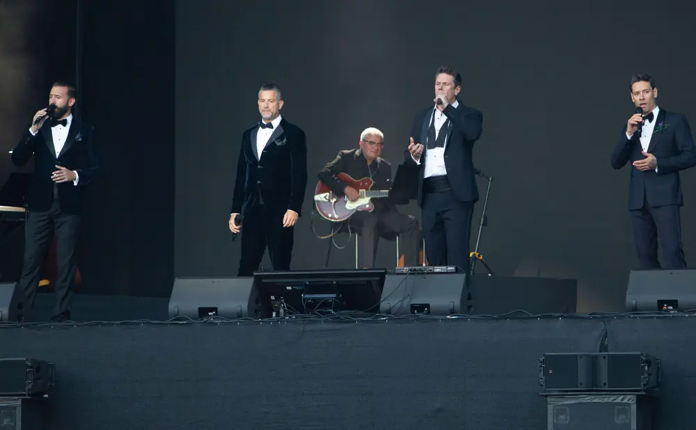 Five men in tuxedos perform on stage with microphones and a guitar.