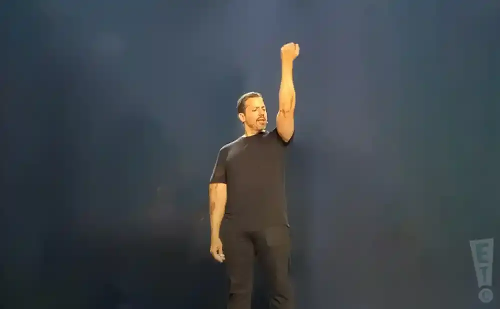A man in a black t-shirt and pants stands with his right arm raised in a fist.