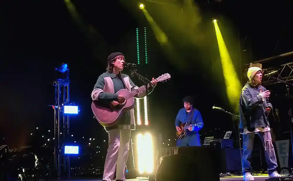 A band performs on stage at night, with bright spotlights and audience lights in the background.