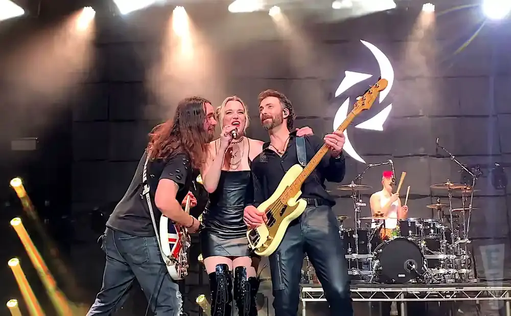 A rock band performs on stage with bright lights and a backdrop featuring a circular logo.