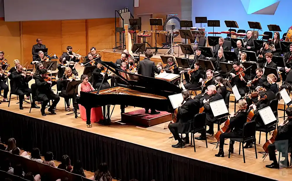 A pianist in a pink dress plays a grand piano with a symphony orchestra behind her.