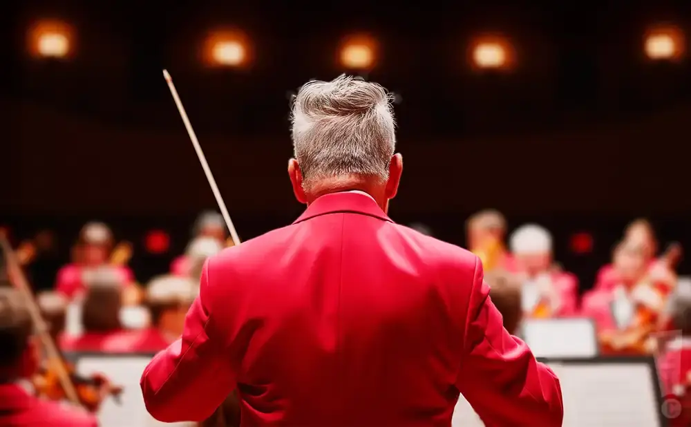 Conductor in a red jacket leading an orchestra in front of blurred audience.