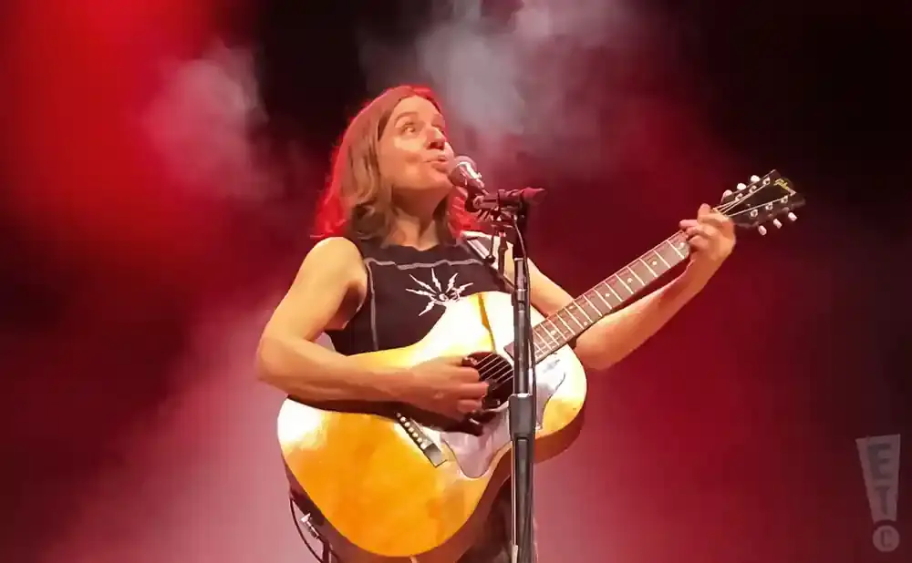 A woman plays an acoustic guitar on stage under red lights.