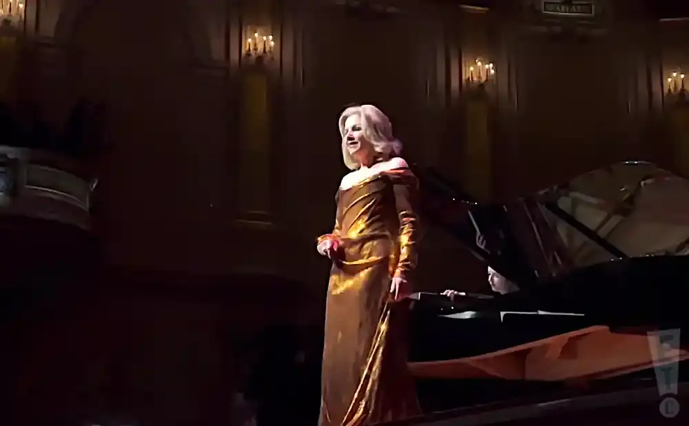 A woman in a golden gown sings on stage next to a grand piano.