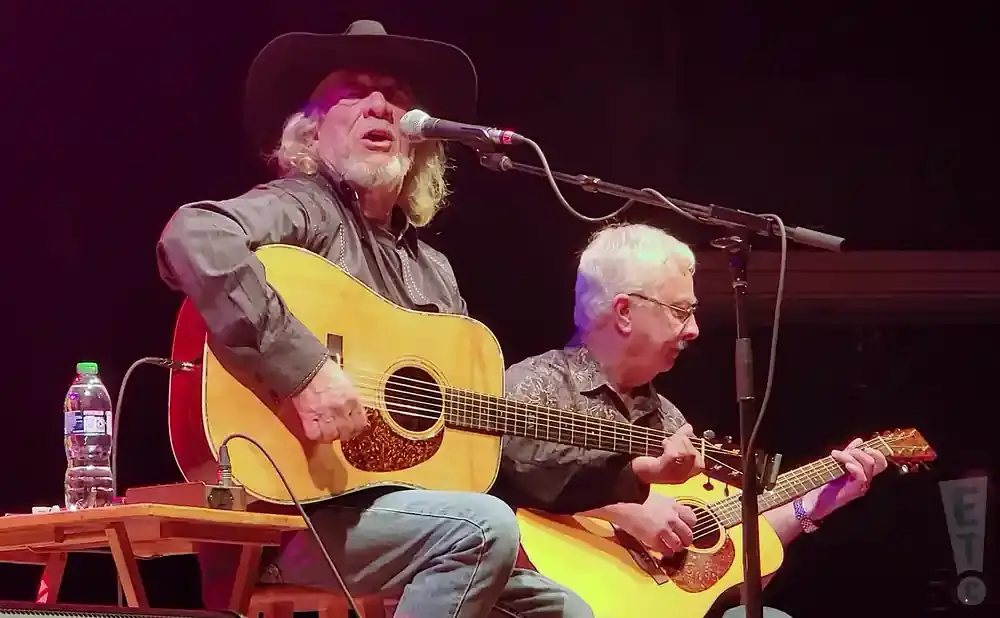 Musicians on stage playing acoustic guitars; one with a cowboy hat sings into a microphone.