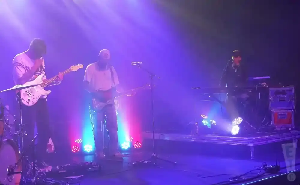 A band performs on stage with purple and blue lighting. Two guitarists and a keyboardist are visible.