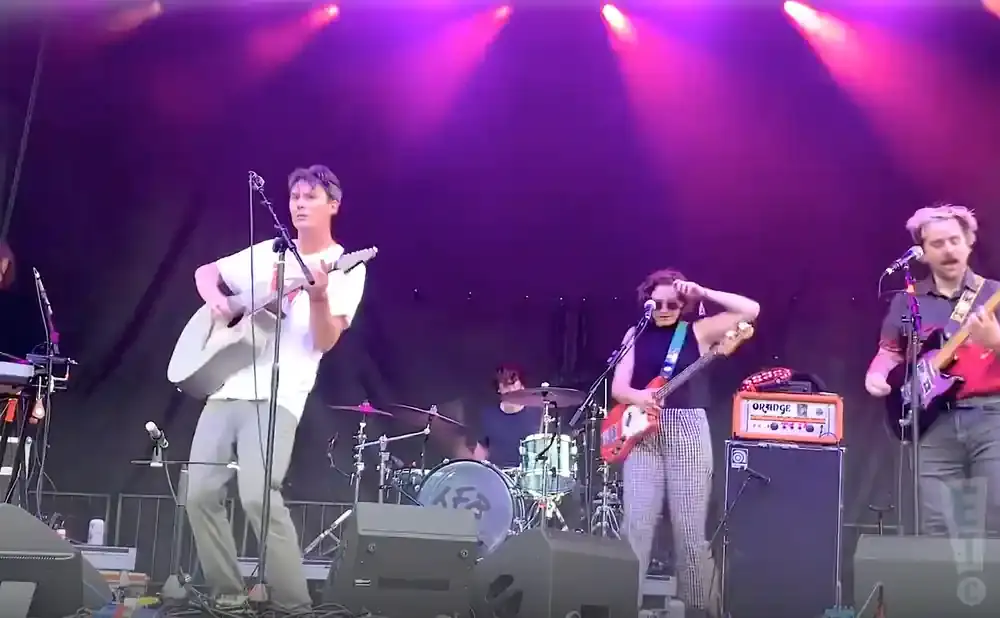 A band performs on stage with purple lighting. A man in a white t-shirt plays an acoustic guitar.