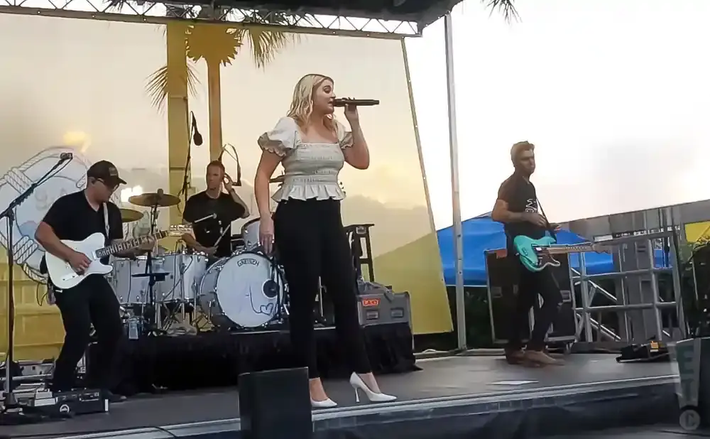 A woman sings into a microphone while playing guitar on stage with her band.