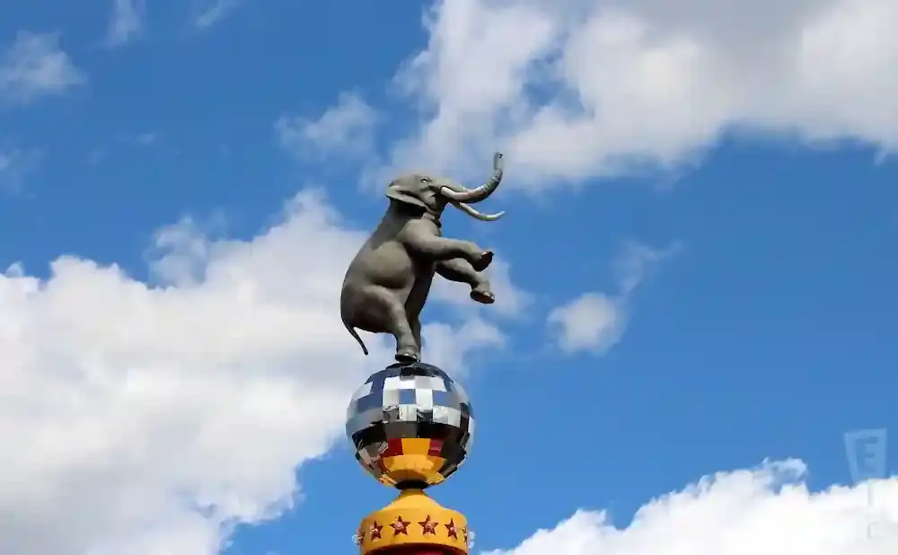 A statue of an elephant balancing on a disco ball against a cloudy blue sky.