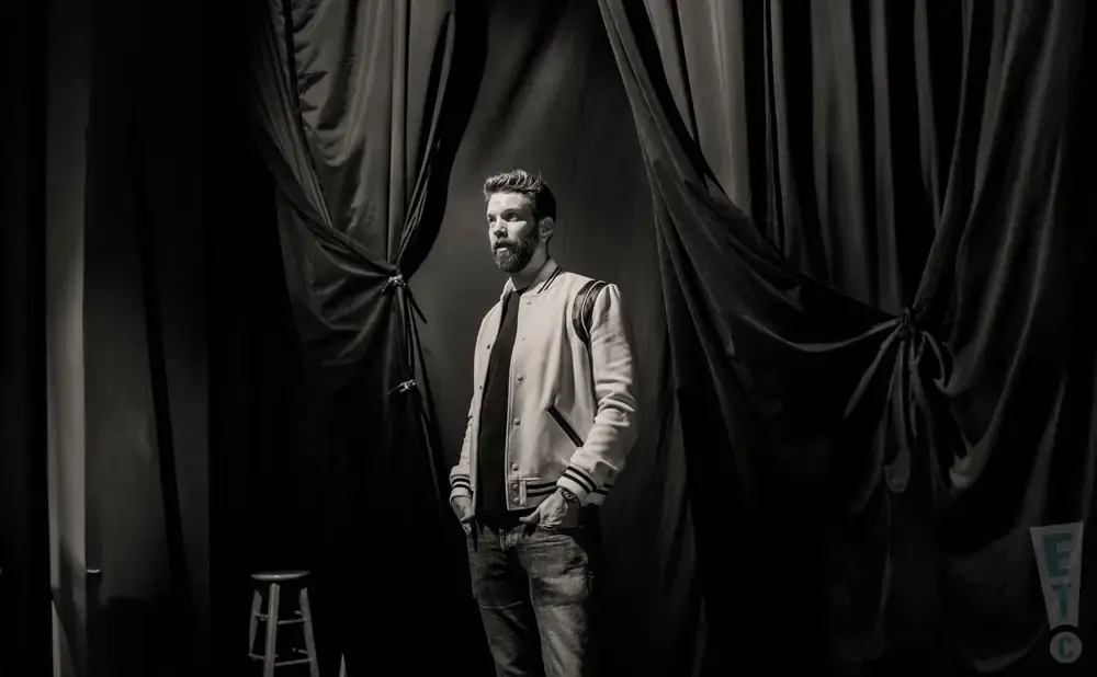 A man with a beard stands on a stage in front of dark curtains, wearing a letterman jacket.