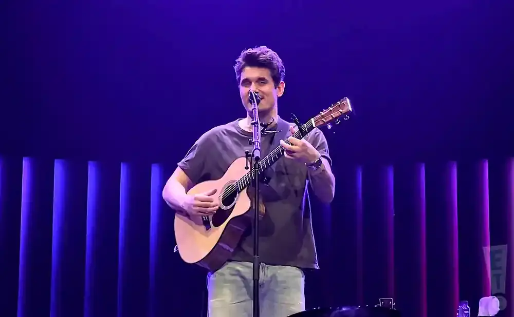 A man with a guitar sings into a microphone on a stage with purple lights.
