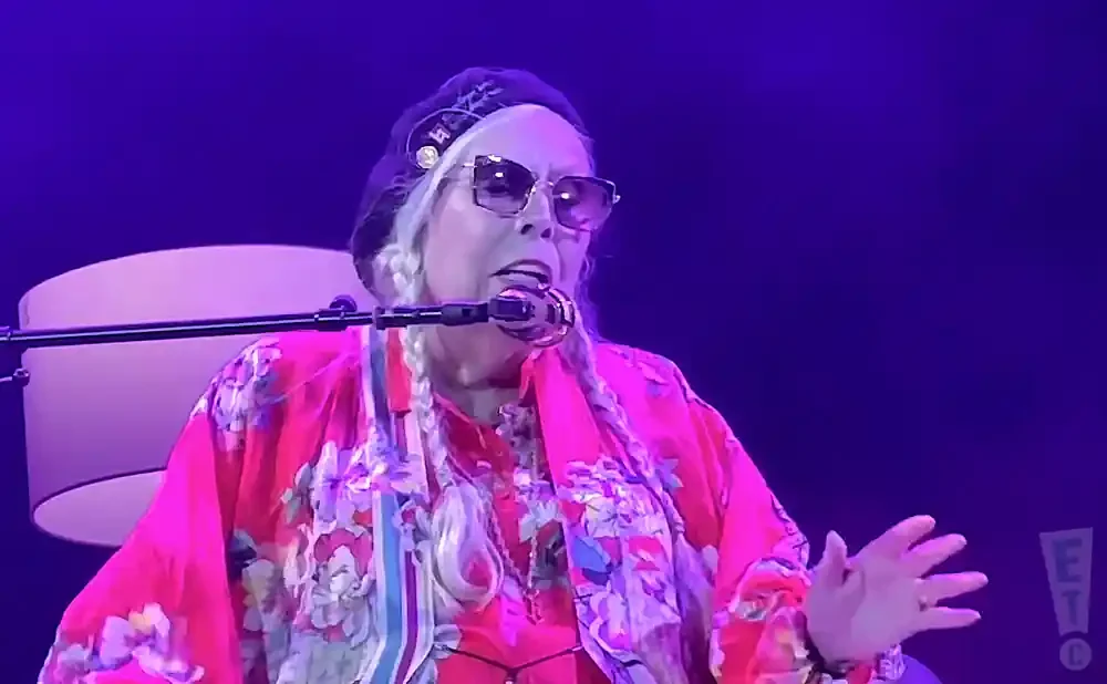 Joni Mitchell sings into a microphone wearing a pink floral kimono and sunglasses.